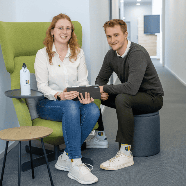 Zwei Personen sitzen auf einem Flur und schauen in die Kamera. Dabei halten Sie ein IPad in der Hand.